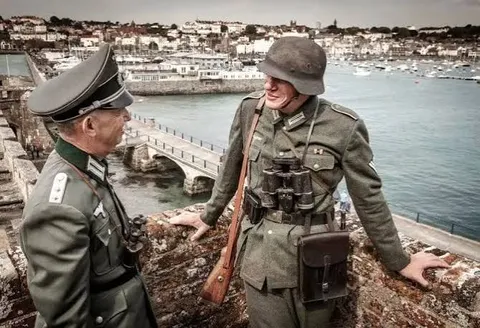 Two German soldiers on a harbour wall above a boat filled harbour and seafront town in occupied Guernsey, Channel Islands