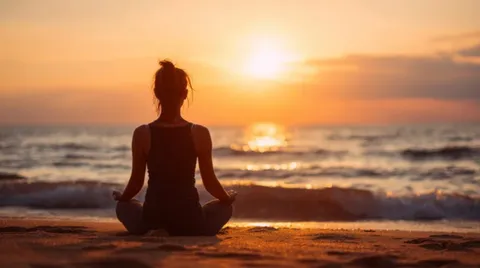A woman sitting cross-legged in meditation on a beach as the sun sets over the sea