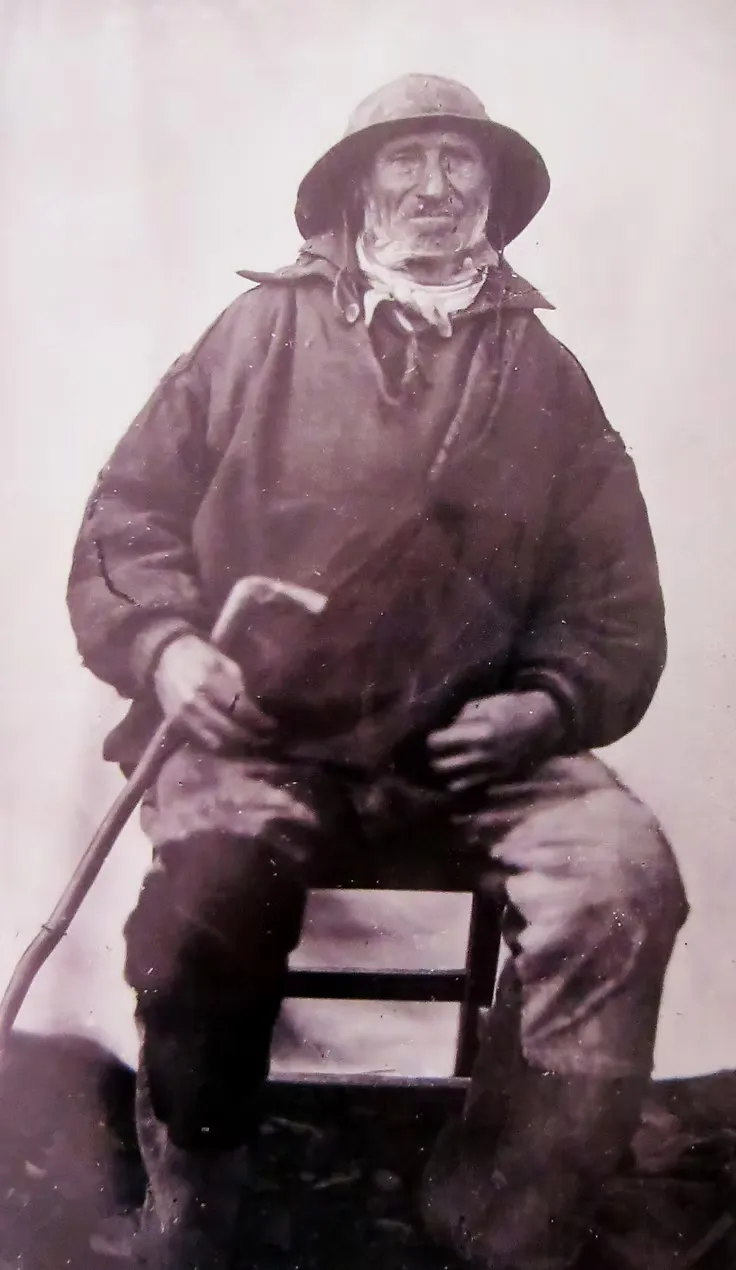 A period photograph of an elderly fisherman in an oilskin smock and hat, seated with a walking stick