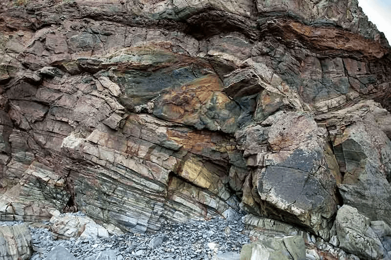 A cliff face of folded and tilted strata in reds, greens and ochres on Sark, Channel Islands