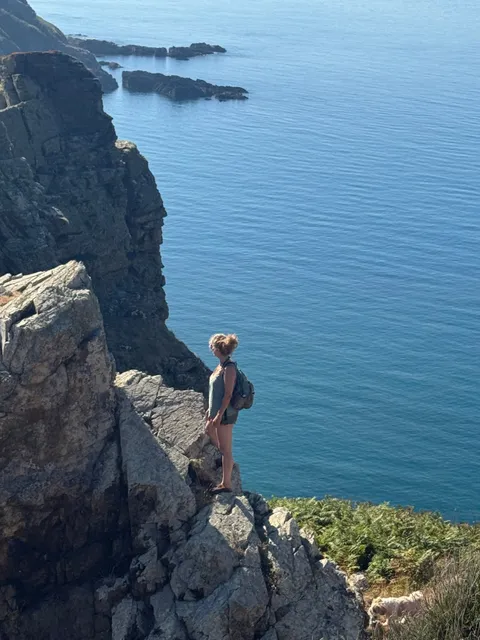 Nadia standing at the edge of a clifftop with a backpack, looking out over the sea on Sark, Channel Islands