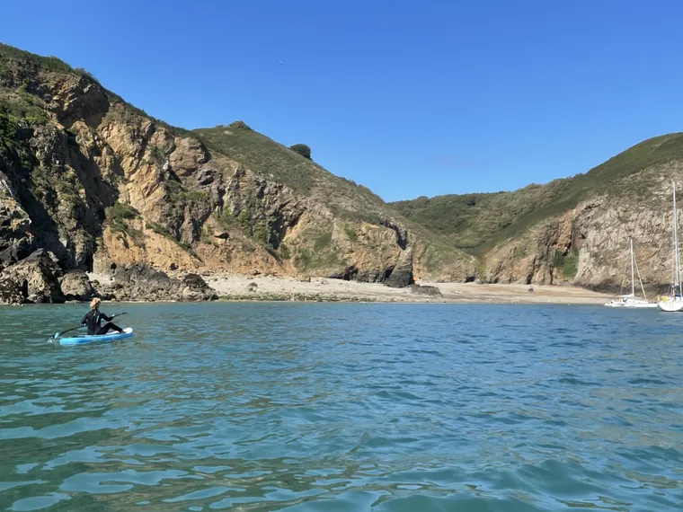 A paddleboarder crossing a clear turquoise bay below the cliffs of Sark