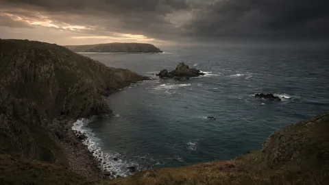 A bay under heavy storm cloud, rock stacks offshore and surf breaking below steep cliffs, the sea the Pilcher party was lost to, Sark, Channel Islands