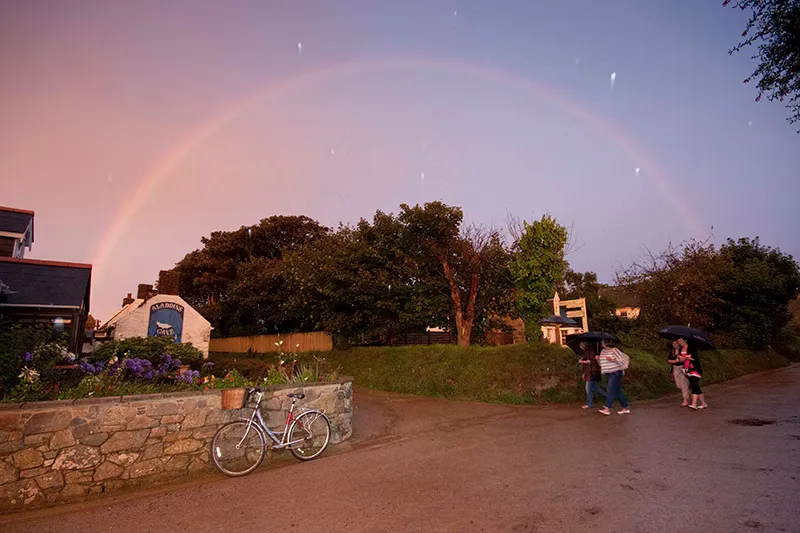 Walkers with umbrellas on a village lane under a rainbow at dusk, Sark, Channel Islands