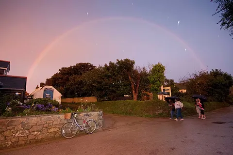 Walkers with umbrellas on a village lane under a rainbow at dusk, Sark, Channel Islands