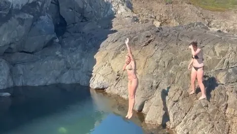 Wild swimming at a deep still rock pool enclosed by pale rock, two women in swimwear on the rocks above the water, Sark, Channel Islands